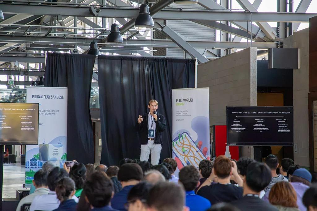 Eoin O'Doherty speaking on stage at Plug and Play San Jose, holding a microphone, with a presentation slide behind him and the audience in front